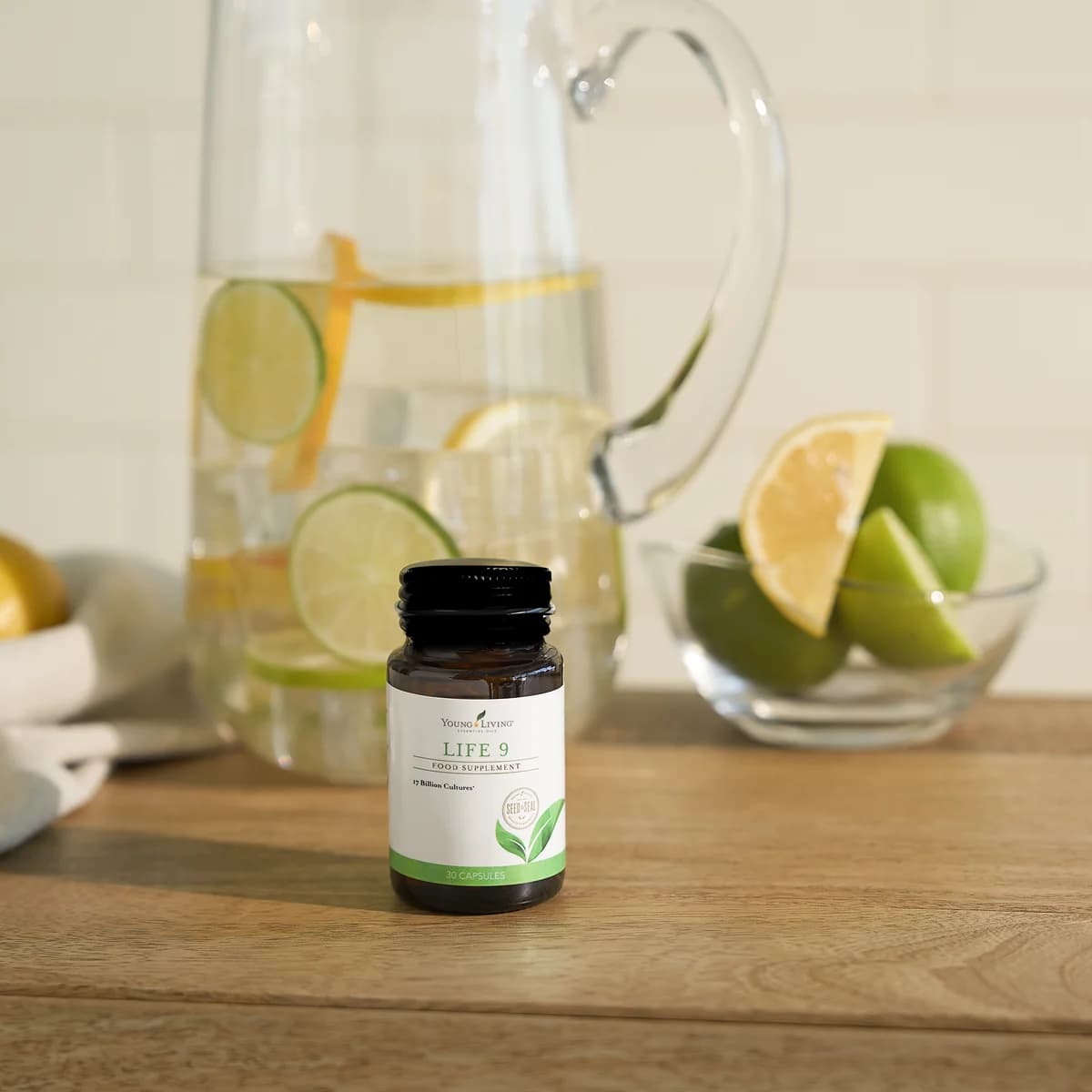 Eine Kapseldose Young Living Life 9 steht auf einer Holzplatte vor einem Glaskrug mit Zitronenwasser und einer Glasschale mit Limetten und Zitrone.
