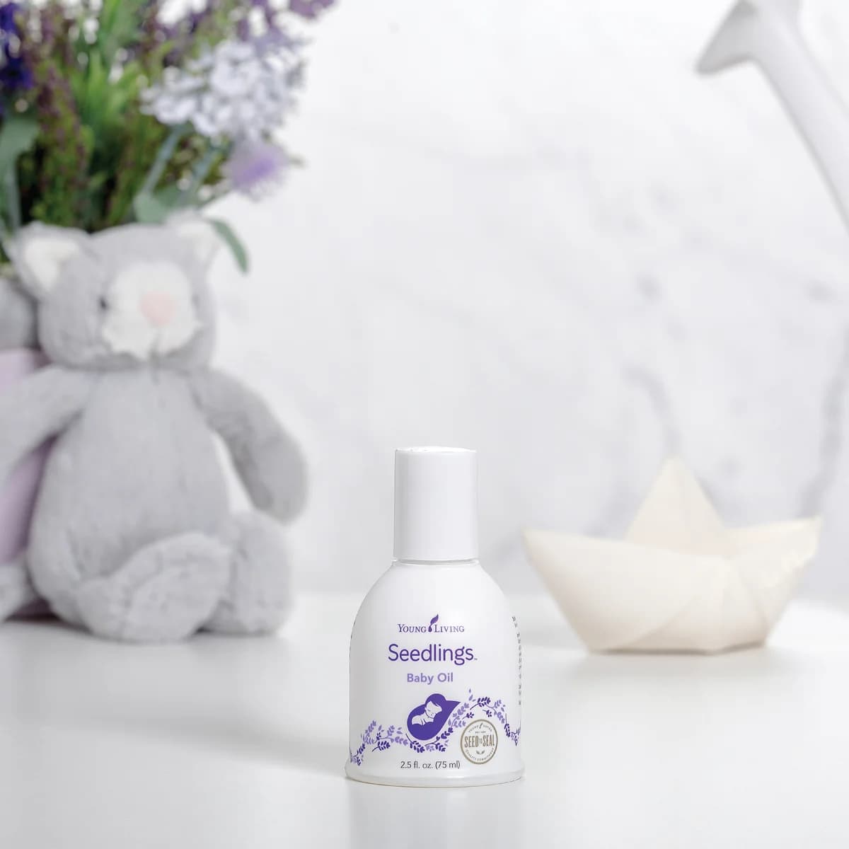 Eine Flasche Young Living Seedlings Baby Oil 75 ml steht auf einer weißen Fläche neben einem grauen Plüschtier, dahinter Lavendel und ein Papierboot.