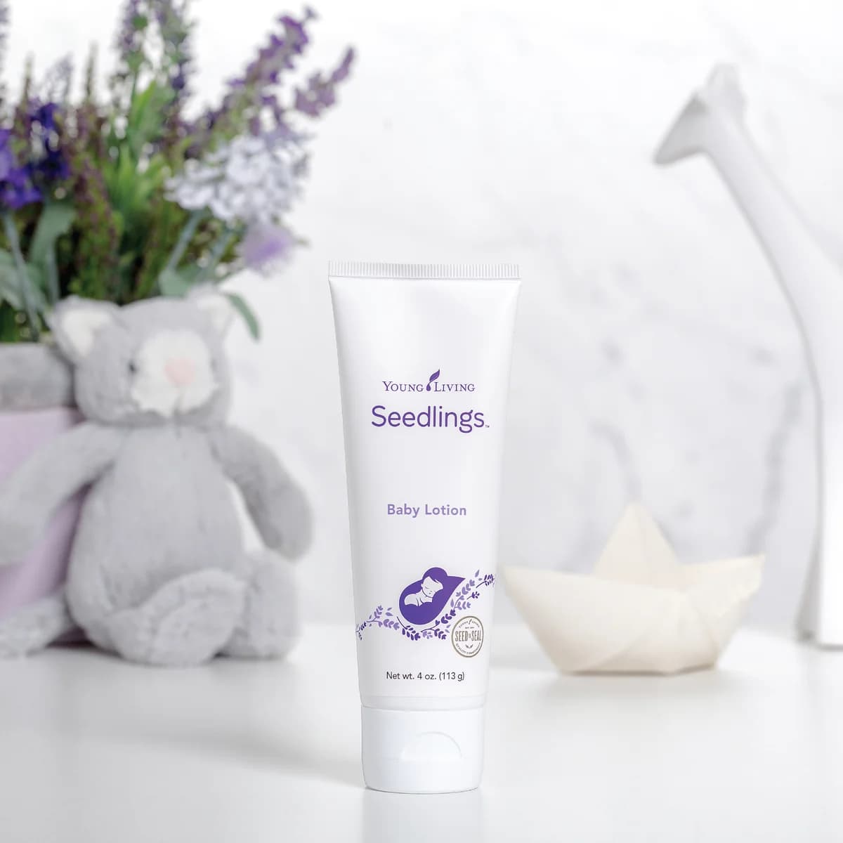 Eine Tube Young Living Seedlings Baby Lotion 113 g steht aufrecht neben einem grauen Plüschtier, dahinter Lavendel, ein Papierboot und eine weiße Giraffenfigur.