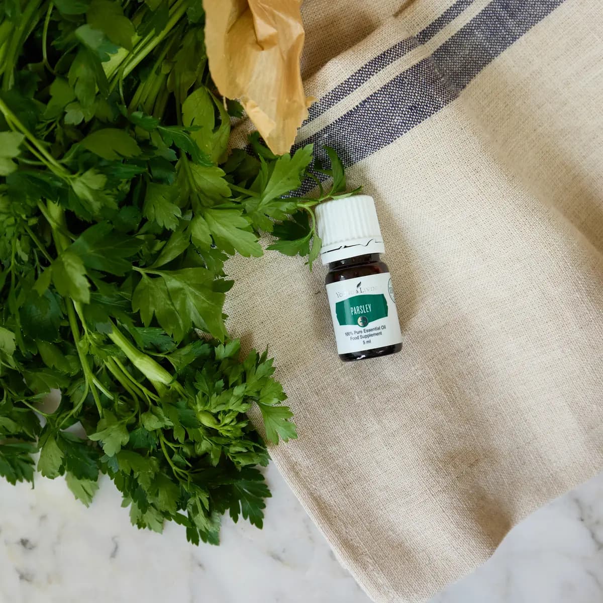 Ein Fläschchen Young Living Parsley+ 5 ml liegt auf einem hellen Leinentuch neben einem Bund frischer Petersilie.