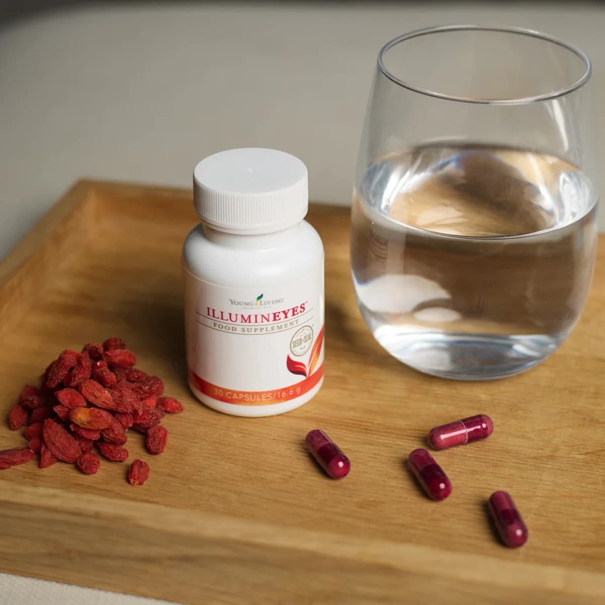 Eine Kapseldose Young Living IlluminEyes steht auf einem Holztablett neben roten Trockenbeeren und einem Glas Wasser.