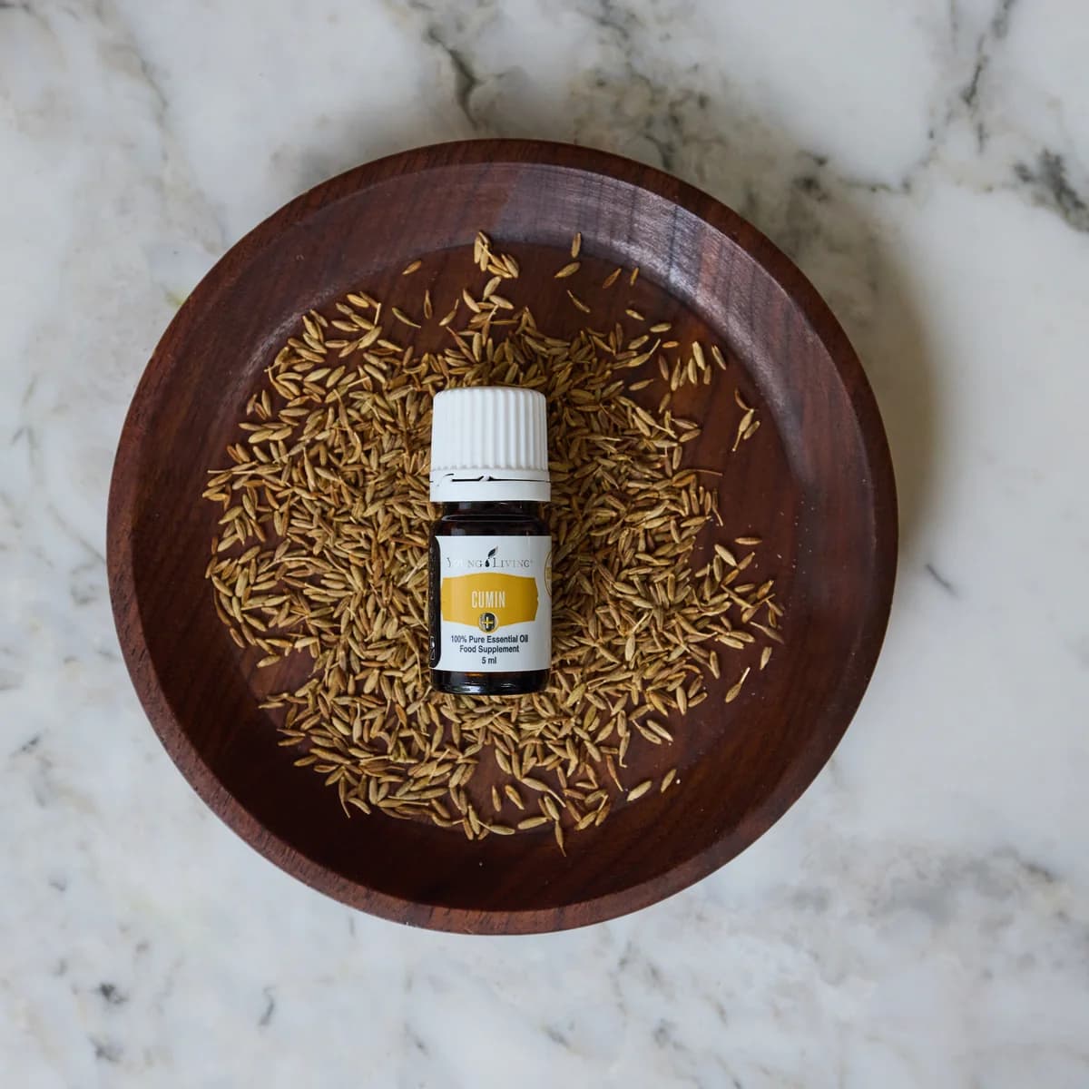 Ein Fläschchen Young Living Cumin+ 5 ml steht in einer dunklen Holzschale voller Kreuzkümmelsamen auf einer Marmorplatte, von oben fotografiert.