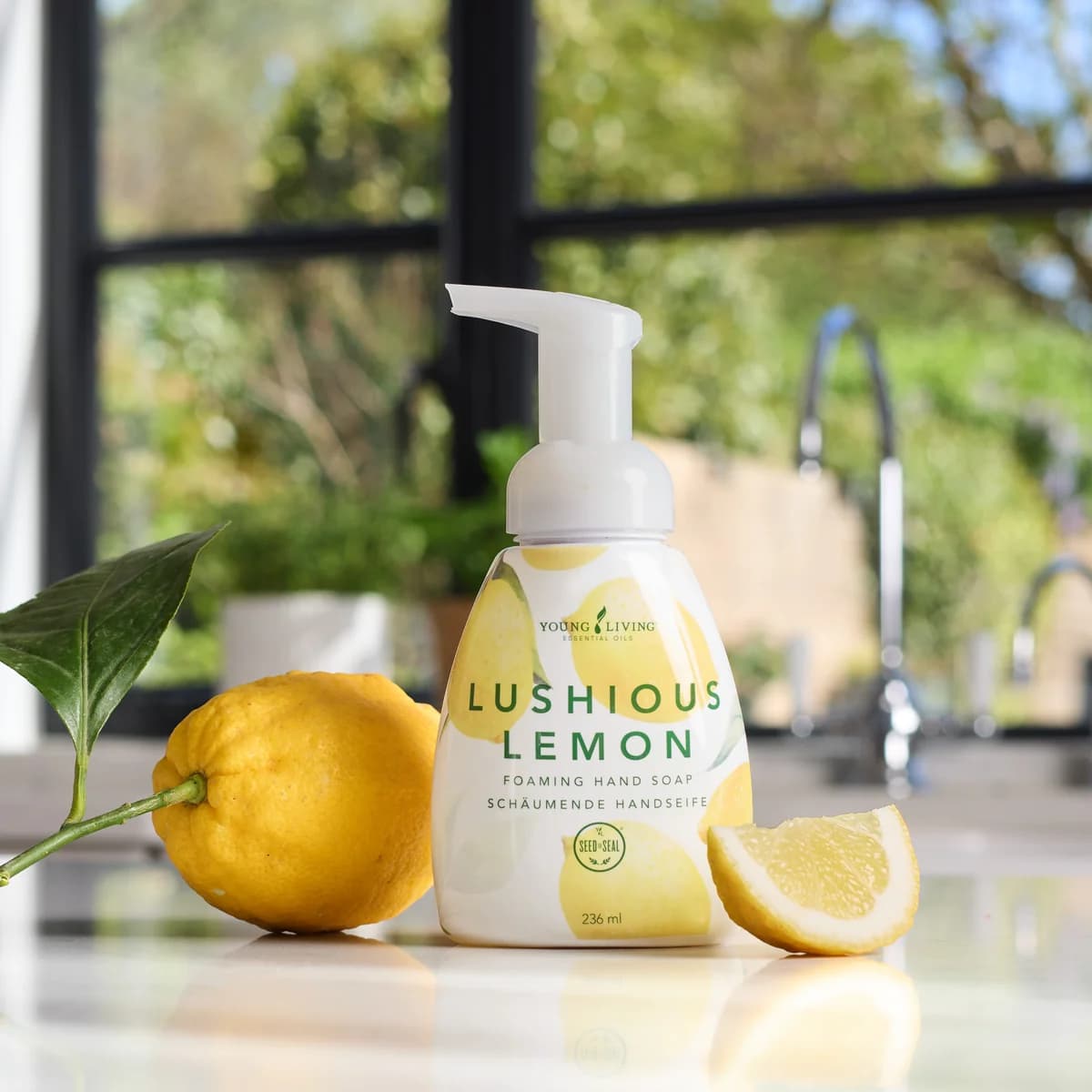 Eine Pumpflasche Young Living Lushious Lemon Foaming Hand Soap steht auf einer weißen Küchenzeile neben einer Zitrone und einem Zitronenstück vor einem Fenster.