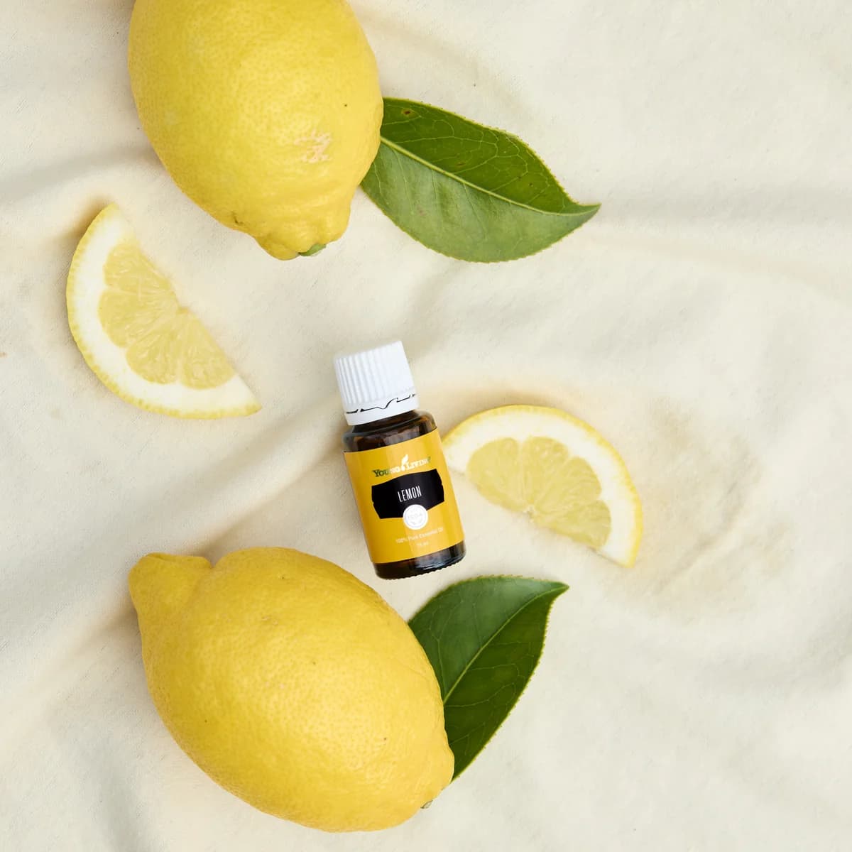 Ein Fläschchen Young Living Zitrone 15 ml liegt auf hellem Stoff zwischen zwei ganzen Zitronen, zwei Zitronenspalten und grünen Blättern, von oben fotografiert.