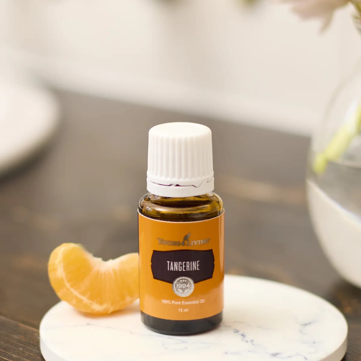 Ein Fläschchen Young Living Mandarinenöl 15 ml steht auf einem runden Marmoruntersetzer neben einer Mandarinenspalte, dahinter unscharf ein Blumenstrauß.
