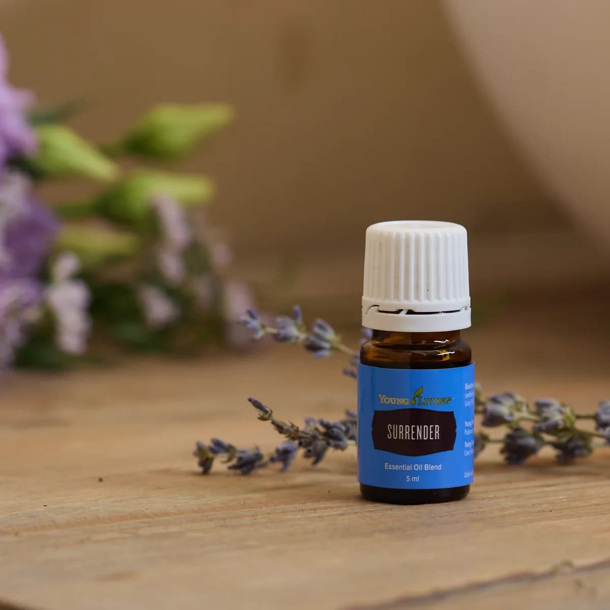 Ein Fläschchen Young Living Surrender 5 ml steht auf verwittertem Holz zwischen Lavendelzweigen, im Hintergrund unscharf lila Blüten und eine weiße Schale.