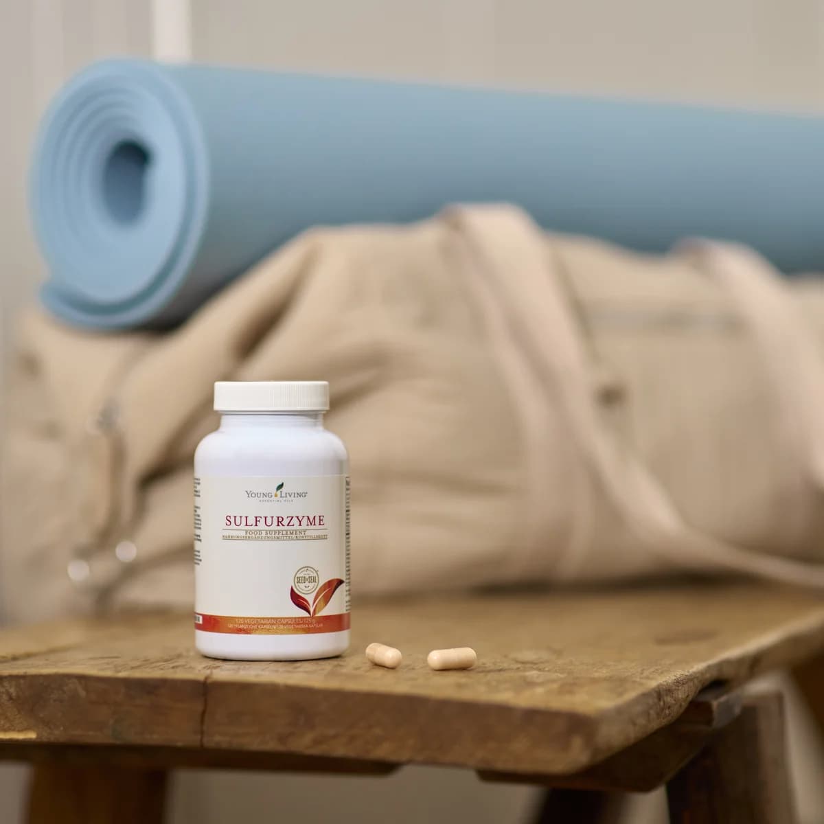 Eine weiße Dose Young Living Sulfurzyme steht auf einer Holzbank neben zwei Kapseln, im Hintergrund eine Yogamatte und eine Sporttasche.