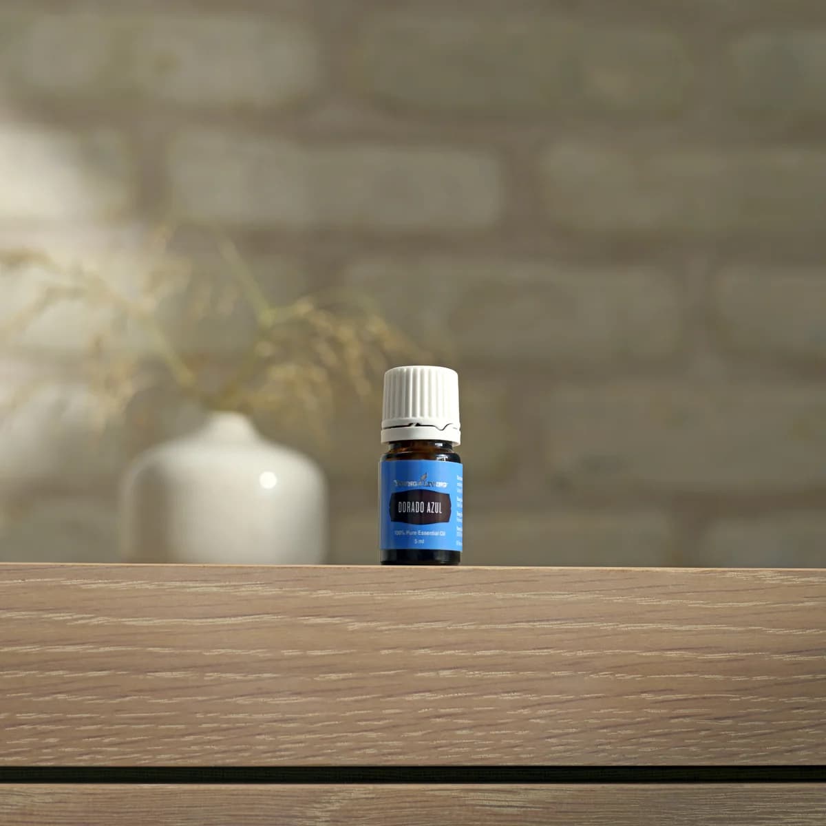 Ein Fläschchen Young Living Dorado Azul Öl 5 ml steht auf einem hellen Holzsideboard vor einer weißen Backsteinwand neben einer hellen Vase.