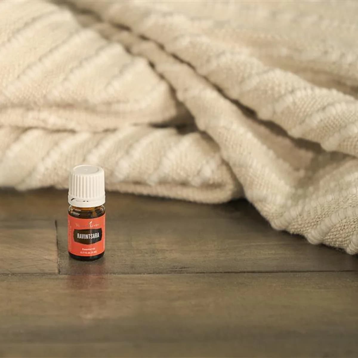 Ein Fläschchen Young Living Ravintsara Öl 5 ml steht auf einem dunklen Holzboden vor einer gefalteten cremefarbenen Strickdecke.