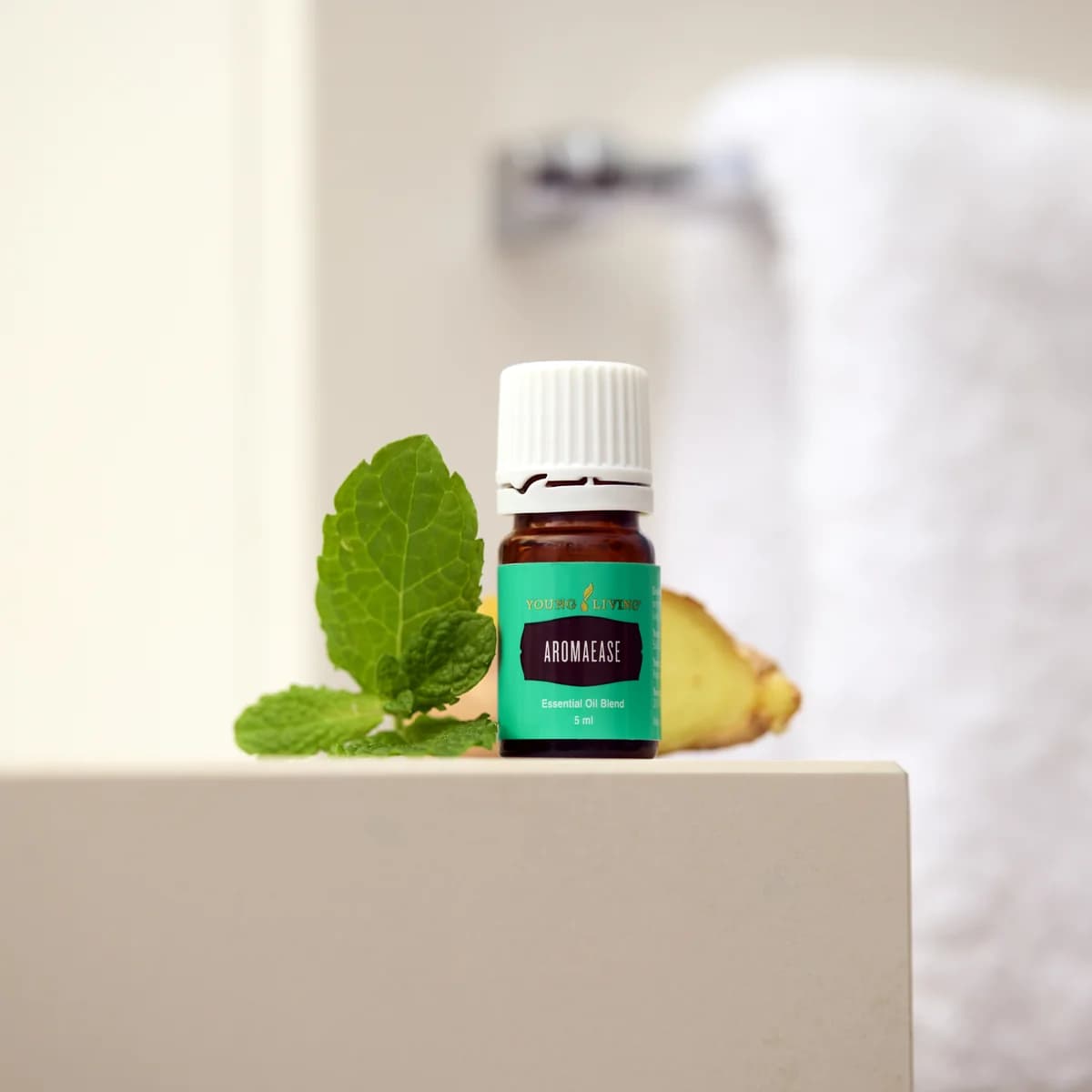 Ein Fläschchen Young Living AromaEase 5 ml steht auf einer weißen Kante neben Minzblättern und einem Stück Ingwer, dahinter ein weißes Handtuch.