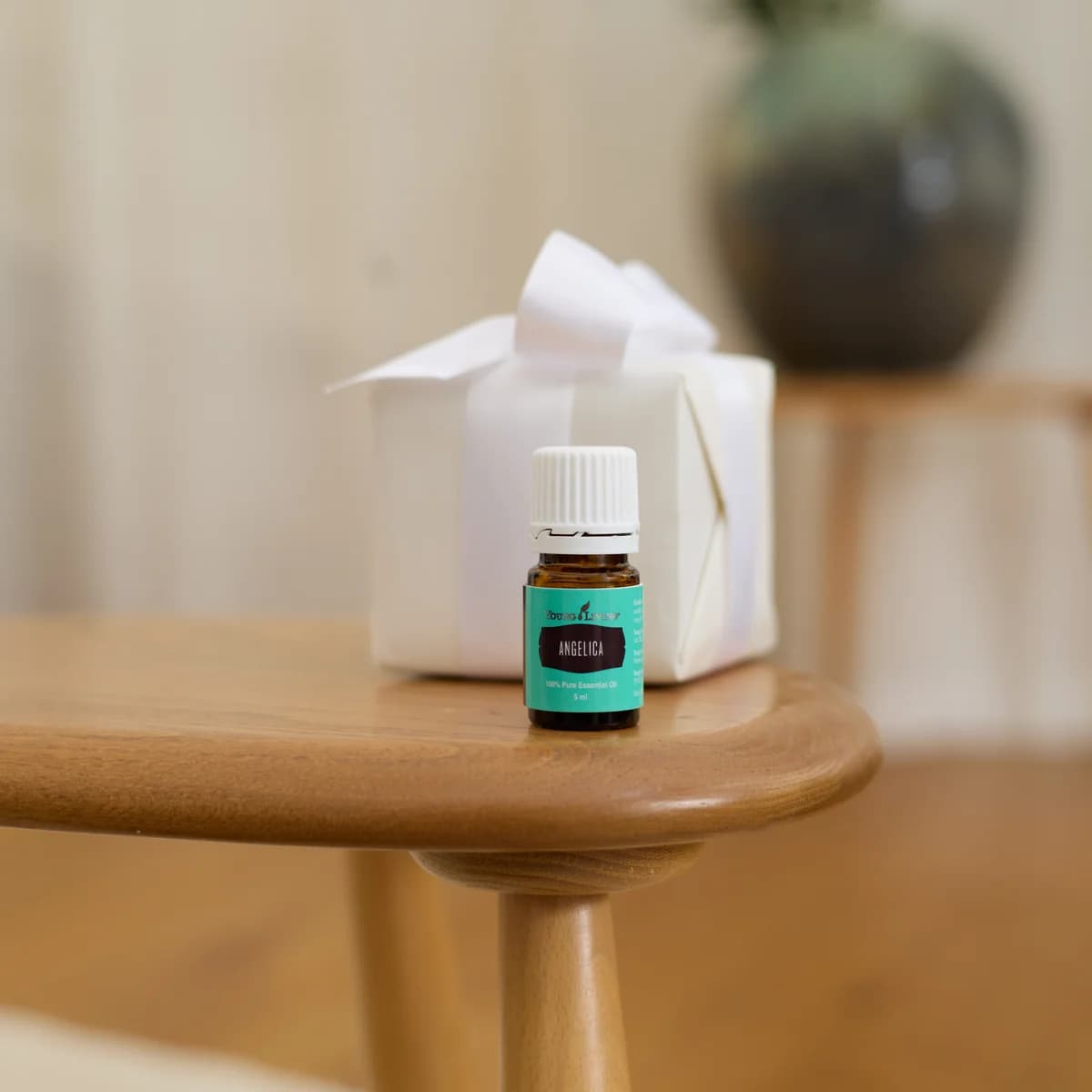 Ein Fläschchen Young Living Engelwurzöl 5 ml steht auf einem hellen Holztisch vor einem weiß verpackten Geschenk, dahinter ein unscharfer Raum.