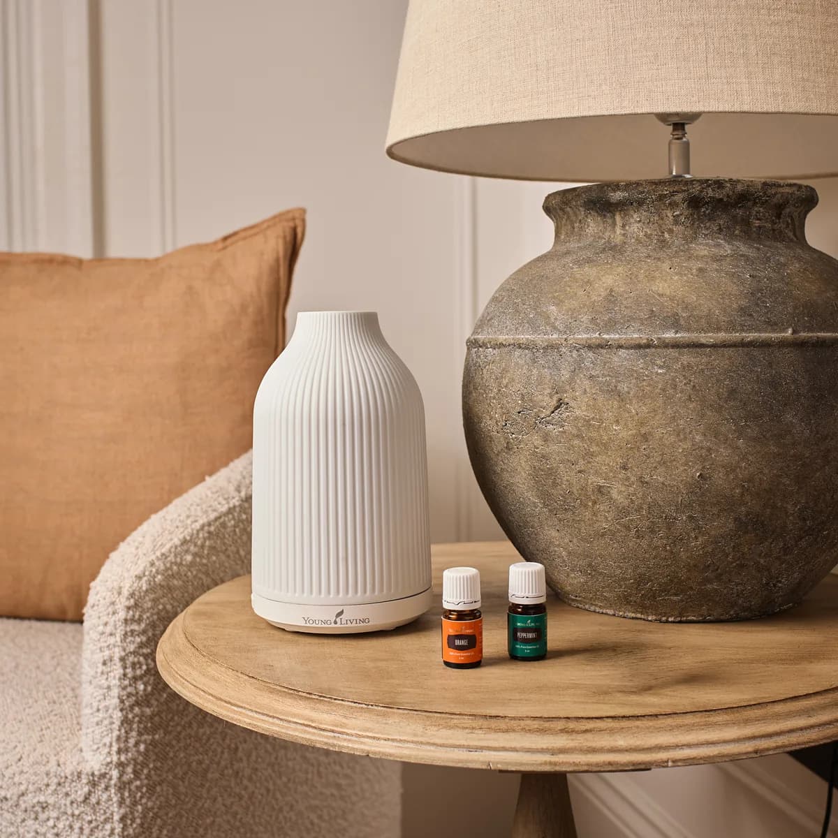 Der weiße Young Living Adobe Mist Ultrasonic Diffuser steht auf einem runden Beistelltisch neben zwei Ölfläschchen und einer großen Steinlampe vor einem Sessel.