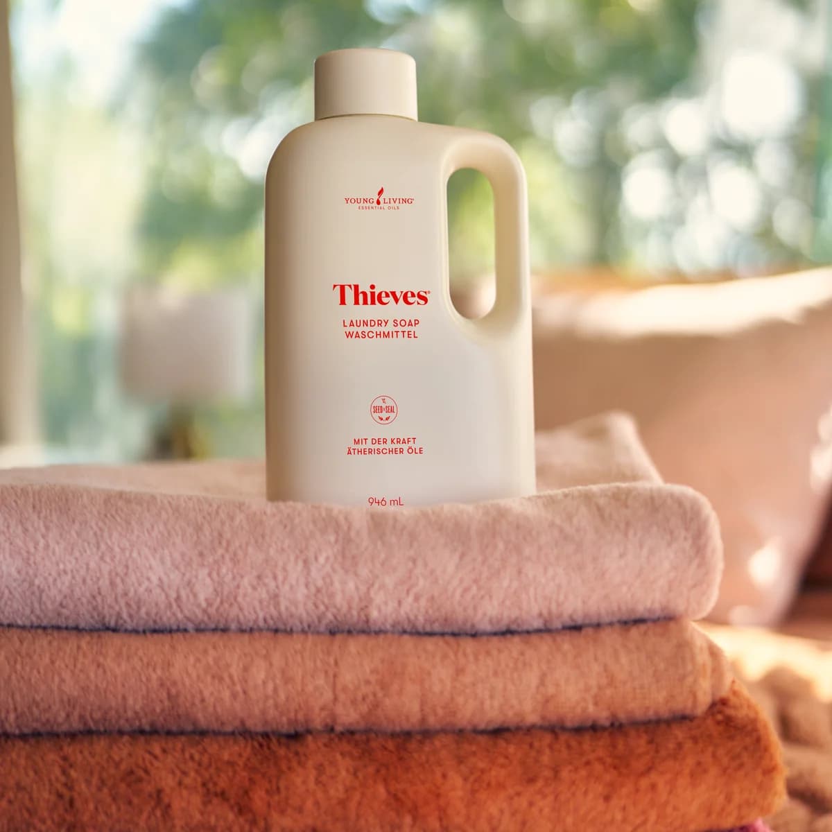 Eine Flasche Young Living Thieves® Laundry Soap 946 ml steht auf einem Stapel gefalteter Handtücher in Rosa, Orange und Pink vor einem hellen Fenster.