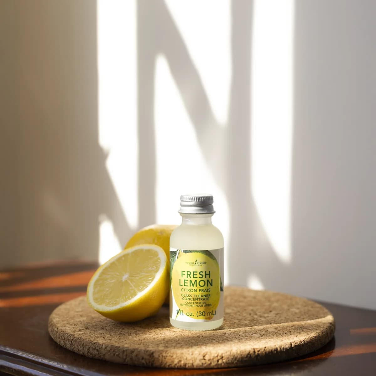 Ein Fläschchen Young Living Fresh Lemon GlassCleaner Concentrate 30 ml steht auf einem Korkuntersetzer neben einer halbierten Zitrone im Sonnenlicht.