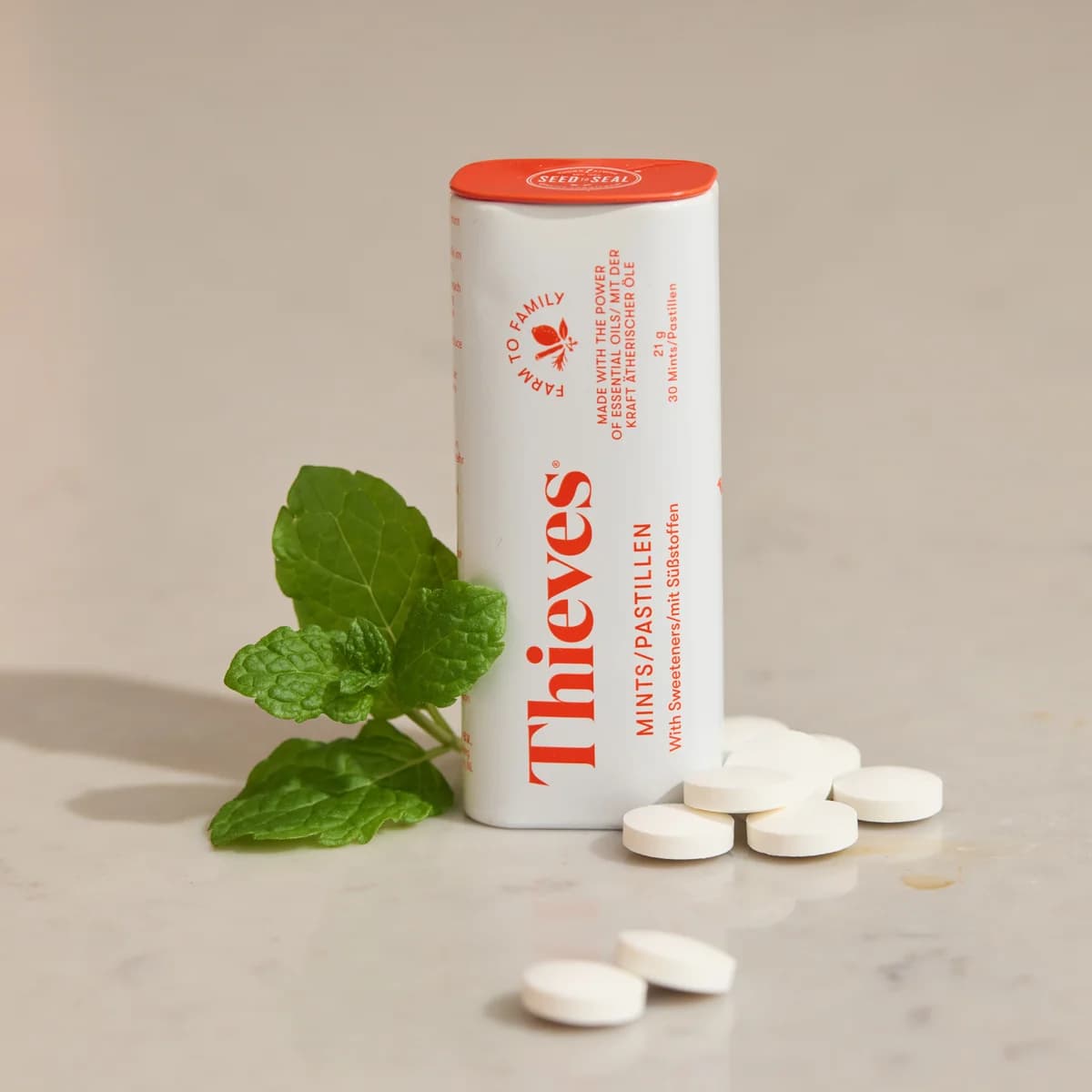 Ein weißer Spender Young Living Thieves Mints steht auf heller Steinfläche, davor liegen einige weiße Pastillen und zwei frische Minzblätter.