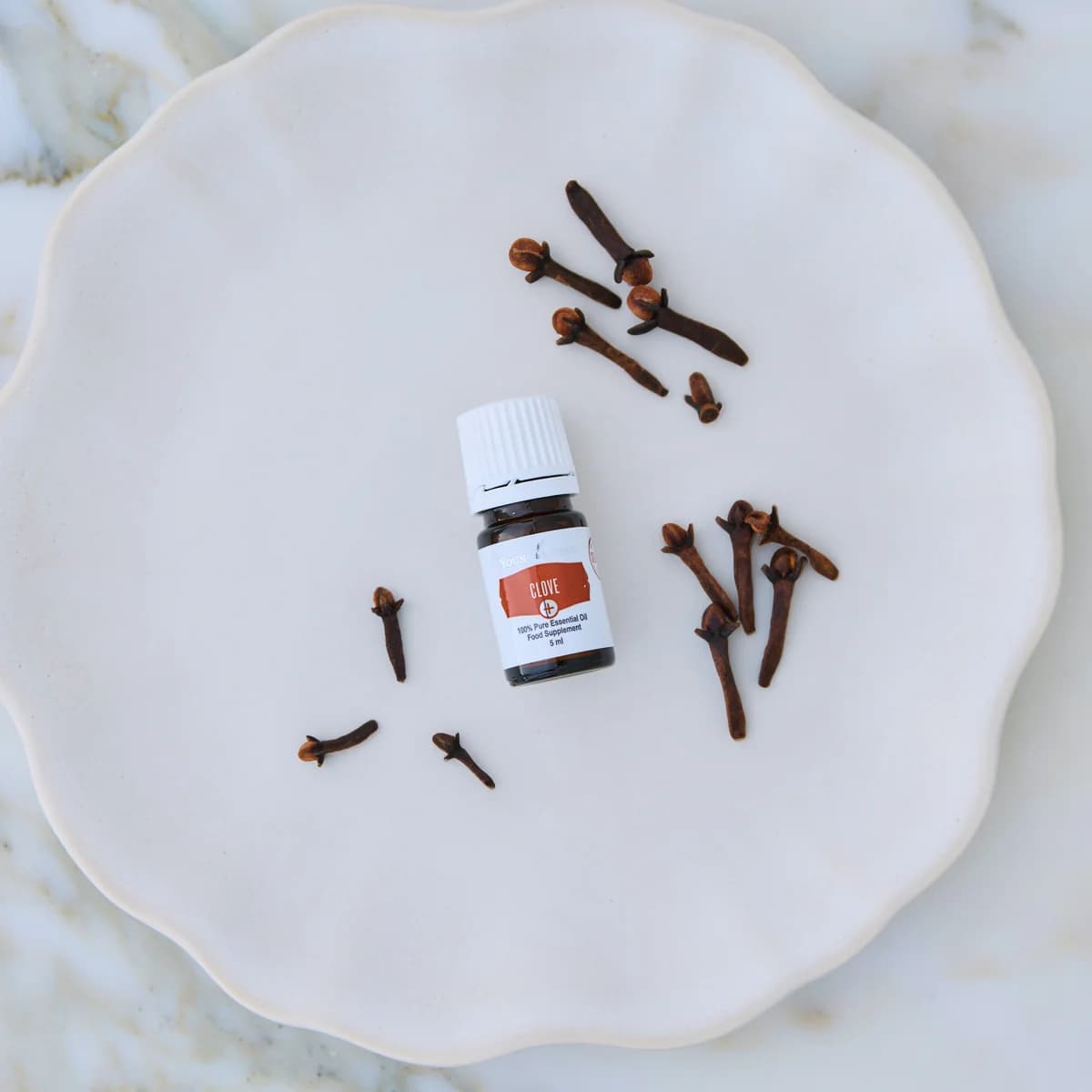 Ein Fläschchen Young Living Clove+ 5 ml liegt auf einem weißen Keramikteller zwischen verstreuten Gewürznelken.