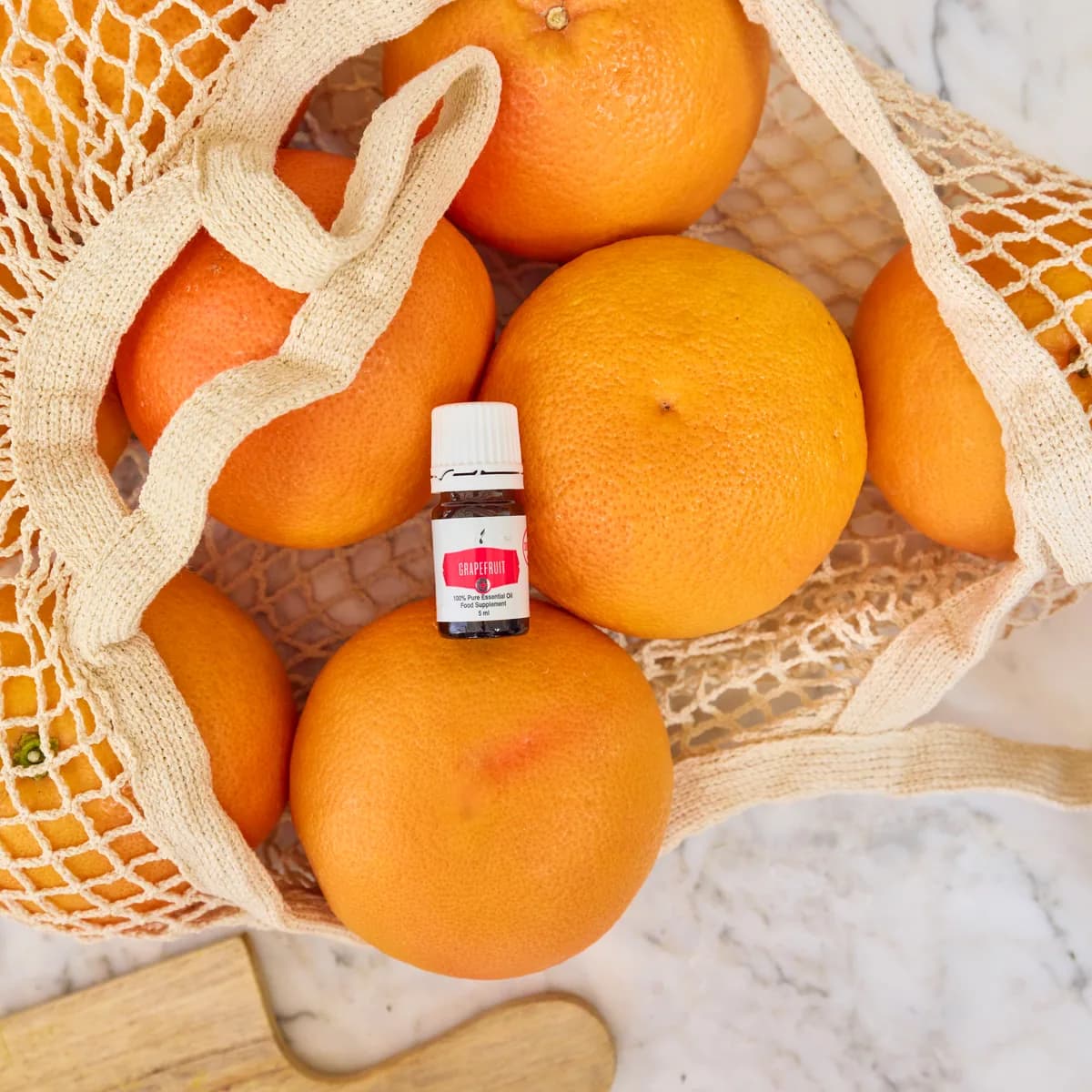 Ein Fläschchen Young Living Grapefruit+ 5 ml liegt zwischen Grapefruits in einem Netzbeutel aus Baumwolle auf hellem Marmor.