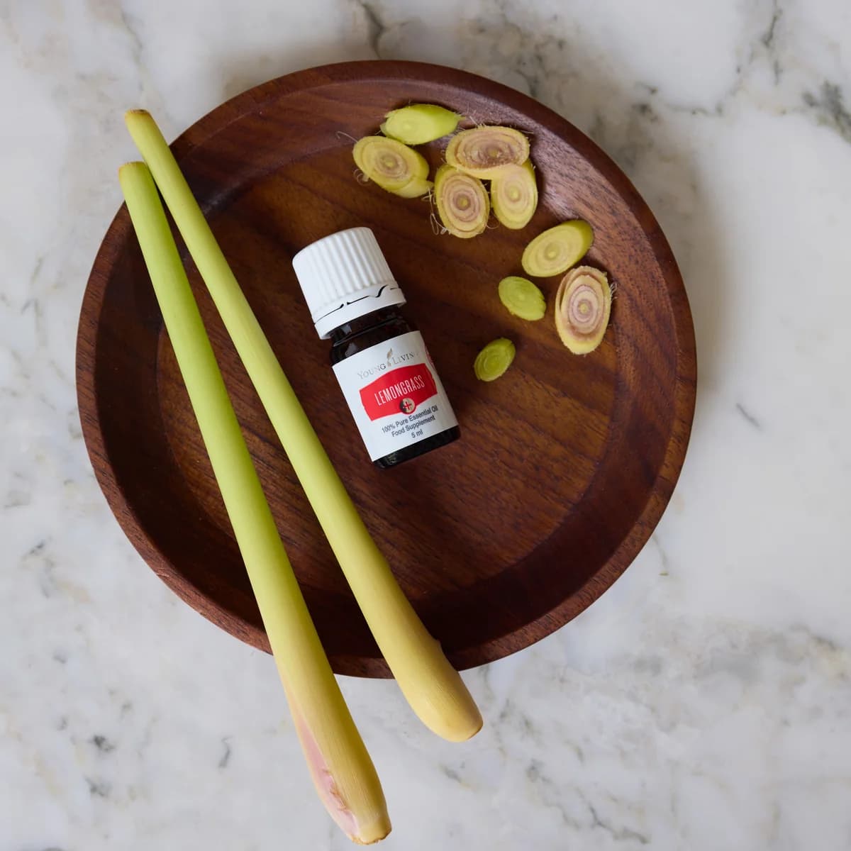 Ein Fläschchen Young Living Lemongrass+ 5 ml liegt in einer dunklen Holzschale neben zwei Zitronengrasstängeln und Zitronengrasscheiben.