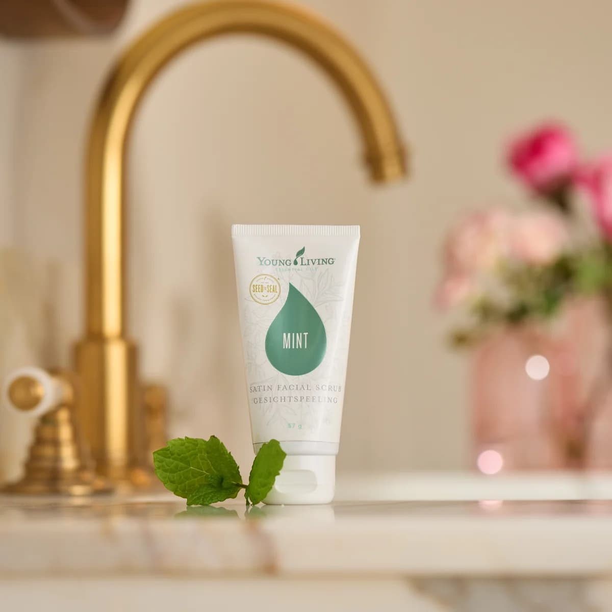Eine Tube Young Living Satin Facial Scrub Mint 57 g steht unter einem goldenen Wasserhahn auf Marmor, davor Minzblätter, dahinter rosa Rosen.