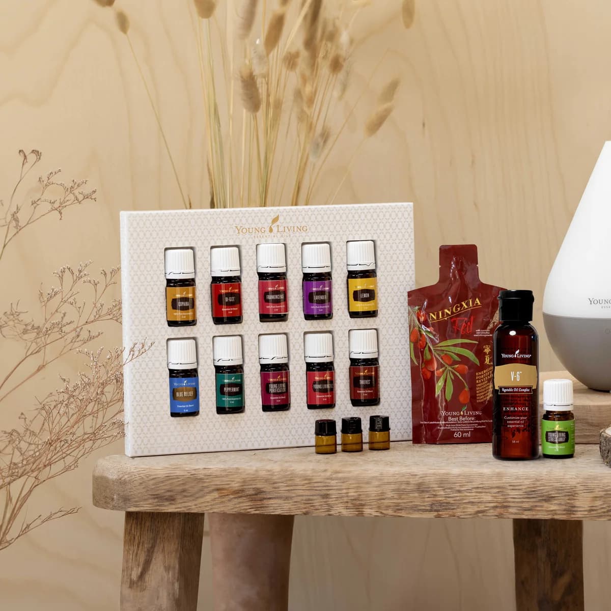 Das Young Living Premium Starter-Kit mit Dewdrop liegt auf einer Holzbank mit zehn Ölfläschchen im Karton, NingXia Red, V-6 und dem Diffuser vor Trockengräsern.