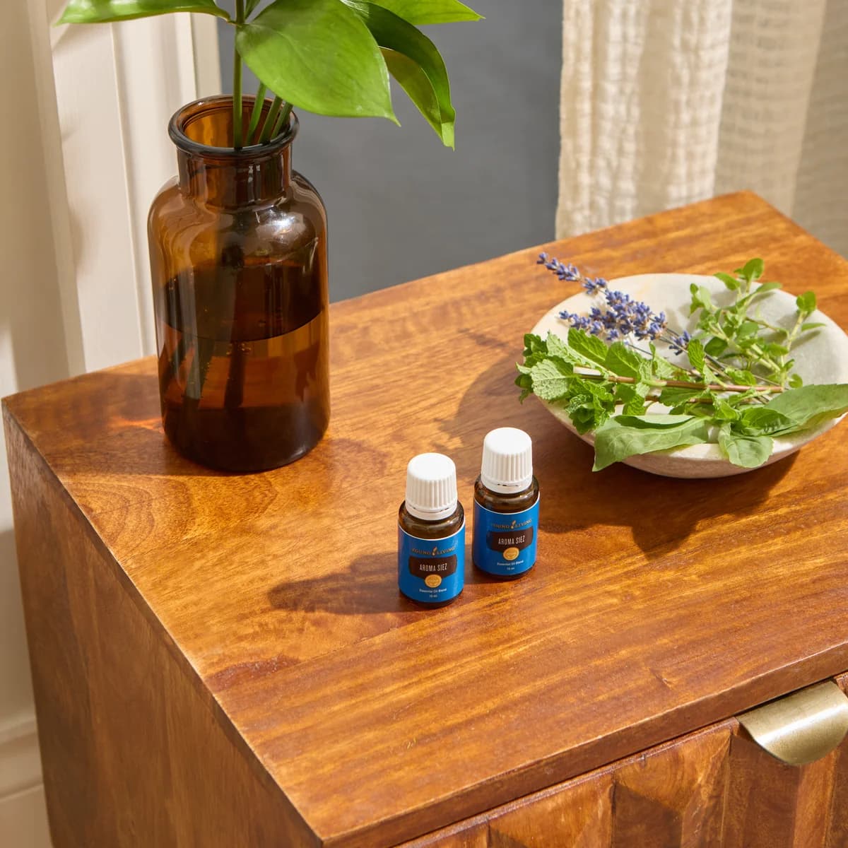 Zwei Fläschchen Young Living Aroma Siez 15 ml stehen auf einer Holzkommode neben einer Braunglasvase und einer Schale mit frischen Kräutern.