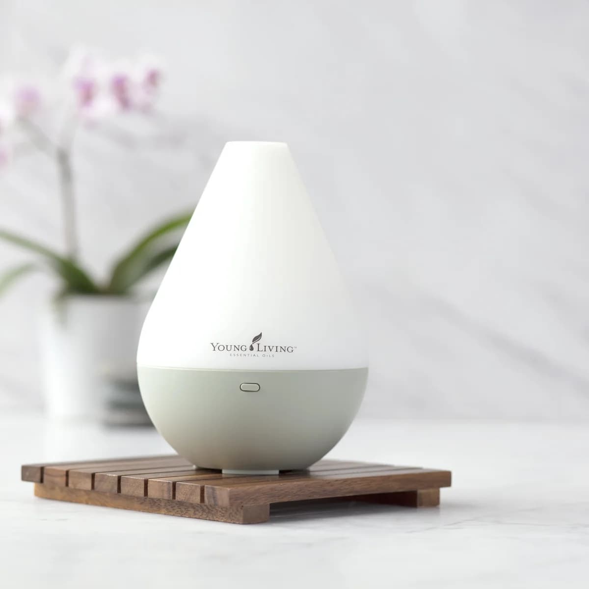 Der weiße Young Living Dewdrop Diffuser steht auf einem Holzrost aus Leisten vor heller Marmorwand, im Hintergrund eine rosa Orchidee.