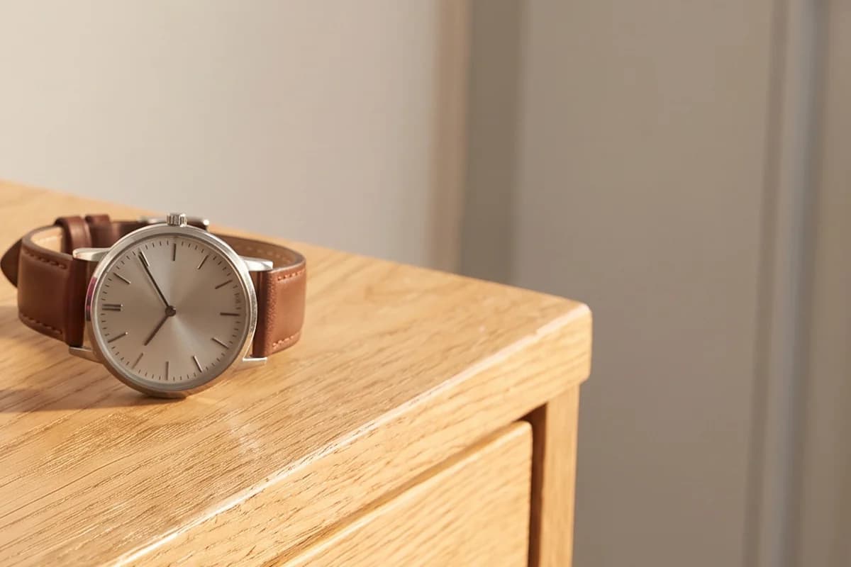 Eine Armbanduhr mit braunem Lederarmband liegt im warmen Licht auf der Ecke einer hellen Holzkommode