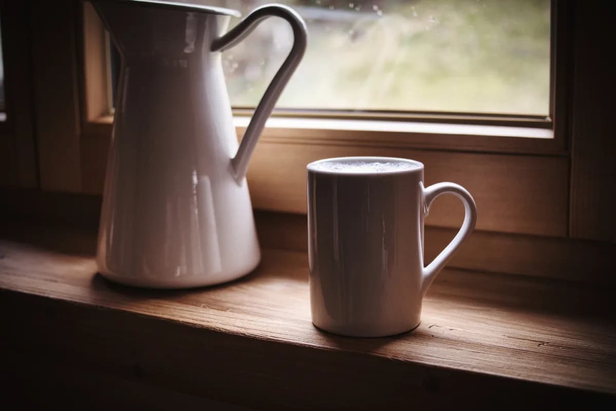 Eine weiße Kanne und eine Tasse auf einer hölzernen Fensterbank im Morgenlicht
